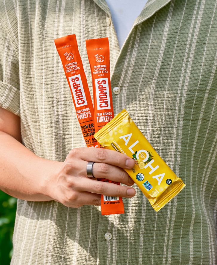 Close-up of a hand holding one protein bar and two jerky sticks.