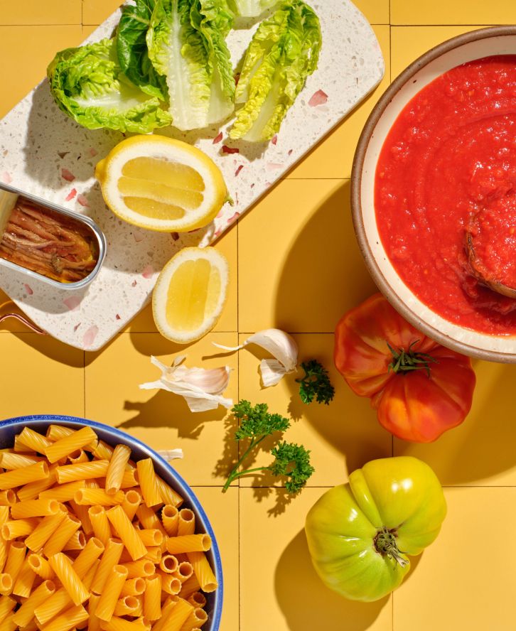 A countertop cooking preparation scene, including pasta, sauce, tomatoes, lettuce, and a lemon.